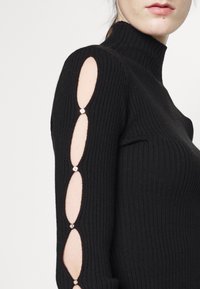 Femme portant un pull à col roulé côtelé noir avec des ouvertures ovales découpées le long de la manche, maintenues par de petites attaches métalliques rondes.