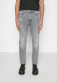Mies, joka pitää yllä slim-fit vaaleanharmaita farkkuja, mustia nauhakengät ja mustaa t-paitaa, seisoo tasaisen valkoista taustaa vasten.