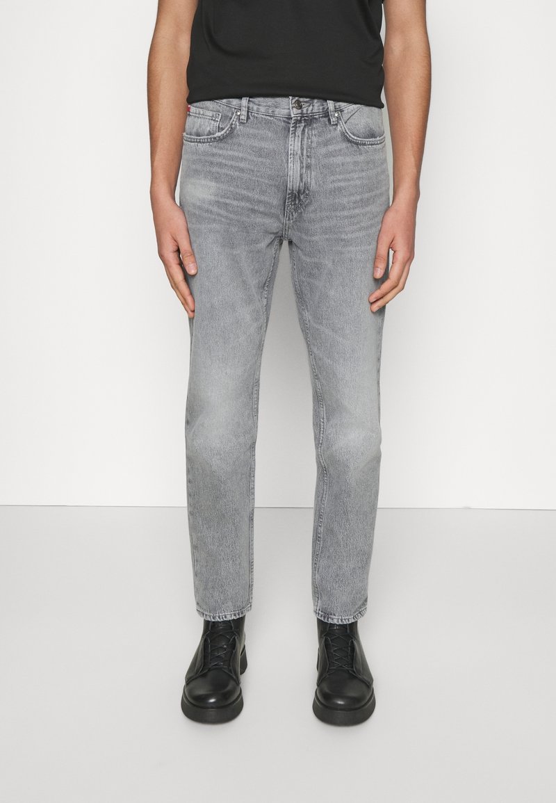 Mies, joka pitää yllä slim-fit vaaleanharmaita farkkuja, mustia nauhakengät ja mustaa t-paitaa, seisoo tasaisen valkoista taustaa vasten.