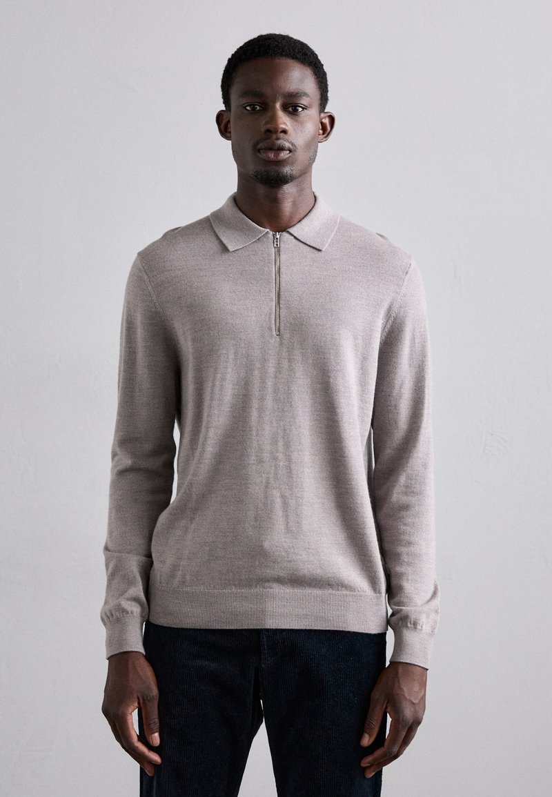 Hellbeige Langarm-Polo-Pullover mit einem halben Reißverschluss, gestrickter Textur und gerippten Bündchen. Kombiniert mit dunkelblauen Jeans.
