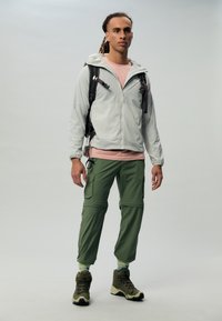 Lichtgrijze zip-up jack, roze T-shirt, groene cargobroek met zakken en bruine wandelschoenen; model staat tegen een neutrale achtergrond.