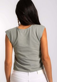 Top sin mangas color verde caqui con cuello redondo, textura suave y sutiles detalles en los hombros, combinado con pantalones blancos.