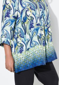Løs-fitting bluse med blå og grøn blomstermønster, prydet med en geometrisk kant nede. Fremstillet af letvægtsstof.