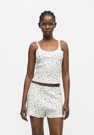 Kvinde iført matchende hvid camisole og shorts med lille blåt blomsterprint mod en ensfarvet lys baggrund.