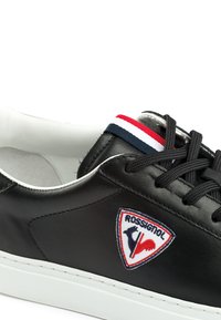 Zwarte leren sneaker met een witte tussenzool, ronde neus, zwarte veters en een patch met een rood, wit en blauw ontwerp aan de zijkant.