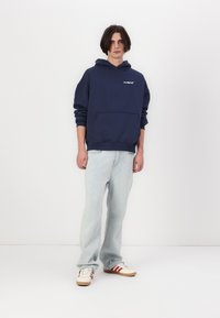Felpa con cappuccio blu navy e tasca frontale, vestibilità rilassata. Abbinato a jeans azzurri e sneakers con strisce rosse. Abbigliamento semplice e informale.