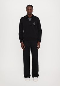 Felpa nera con zip e logo, colletto strutturato e polsini a coste, abbinata a pantaloni neri a gamba larga e sneakers bianche.