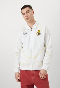 Hvid zip-up jakke med gule syninger, Puma-logo og emblem for Ghana Football Association på brystet. Har elastiske mansjetter og krave.
