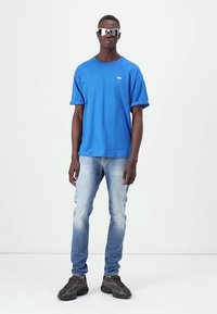 Μπλε βαμβακερό μπλουζάκι, στρογγυλή λαιμόκοψη, κοντό μανίκι; συνδυασμένο με ανοιχτόχρωμο μπλε τζιν και μαύρα αθλητικά παπούτσια. Τετράγωνα γυαλιά ηλίου φοριούνται στο πρόσωπο.