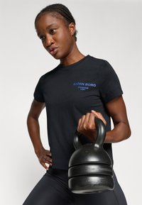 Juodas marškinėliai su mėlynu "BJÖRN BORG" logotipu, dėvimi žmogaus, laikymo juodą kettlebell. Sukirptas su apvalia iškirpte ir trumpomis rankovėmis.