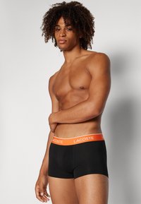 Black boxer briefs with a thick orange waistband featuring the "Lacoste" logo. The fabric appears smooth and stretchy, providing a fitted design.