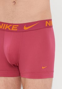 Nike bokserski gaćice u tamnorozoj boji s narančastim elastičnim pojasom, s logotipom brenda i izrađene od glatkog, rastezljivog materijala.