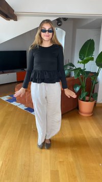 Top peplum negro de manga larga, pantalones de lino gris claro, zapatos planos grises y gafas de sol negras. Ambiente interior con plantas y un sofá.