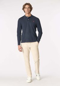 Polo shirt a maniche lunghe blu navy con colletto e logo rosso, abbinato a pantaloni beige slim-fit e sneakers bianche, su uno sfondo neutro.