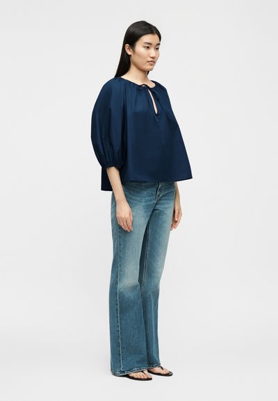 Γυναίκα με μακριά μαύρα μαλλιά, φοράει μια άνετη μπλε μπλούζα με δέσιμο εμπρός και καμπάνα μπλε τζιν, στέκεται μπροστά σε ένα απλό φωτεινό φόντο.