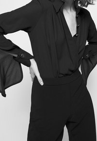 Blusa nera a maniche lunghe con scollatura a V profonda, dettagli con bottoni sul davanti e polsini a campana. Tessuto liscio con una finitura elegante.