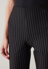 Sorte pinstripe bukser laget av myk stoff, med en tettsittende design, elegante linjer og høy midje. Hånd som hviler på siden.
