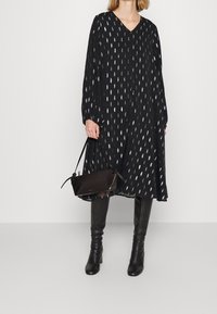 Robe noire à manches longues avec motif en feuilles argentées, décolleté en V et fermeture à boutons, associée à un petit sac à main noir et des bottes noires jusqu'au genou.