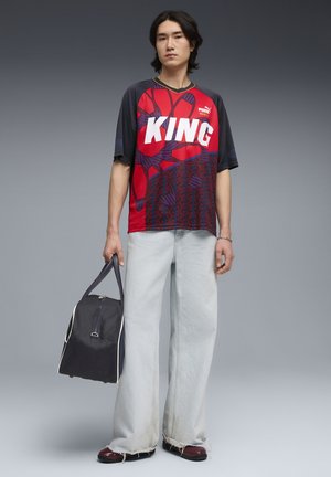 Jeune homme portant un t-shirt rouge et noir "KING" de Puma, un jean large clair, des chaussures bordeaux, tenant un sac de sport noir sur un fond gris dégradé.