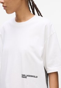Hvid bomulds T-shirt med korte ærmer, der har et sort logo print på siden. Designet inkluderer en kontrasterende grå sideløsning.