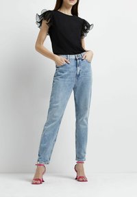 Černý top s průhlednými volánovými rukávy, světle modré džínsy s vysokým pasem a růžové sandály na podpatku. Jednoduchý střih a uvolněný fit.