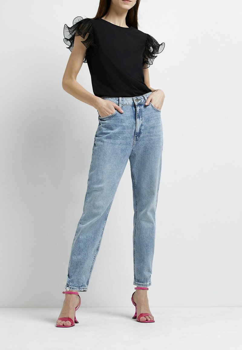 Černý top s průhlednými volánovými rukávy, světle modré džínsy s vysokým pasem a růžové sandály na podpatku. Jednoduchý střih a uvolněný fit.