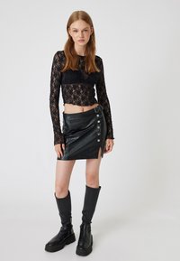 Schwarzes Spitzenshirt mit langen Ärmeln kombiniert mit einem schwarzen Minirock aus Kunstleder mit seitlichen Knöpfen und einem hohen Schlitz bis zum Oberschenkel. Schwarze kniebündige Stiefel.