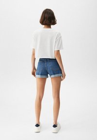 Witte cropped t-shirt, blauwe denim shorts met opgerolde zomen, en witte sneakers met donkerblauwe accenten. Soepele stof en casual pasvorm.