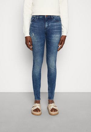 Henkilö, jolla on siniset kapeat farkut, joissa on lievää kulutusta, ja beige käsinkudottu remmisandaalit, seisoo yksinkertaisen vaalean taustan edessä.