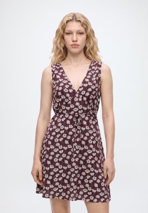 Femme aux cheveux blonds ondulés portant une robe bordeaux sans manches avec des motifs floraux blancs et une ceinture nouée à la taille, debout sur un fond blanc.