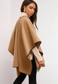 Brūns pončo ar melnu apdari, izgatavots no mīksta auduma. Cieši pārklāj plecus, un tam ir brīvs piegriezums un noapaļoti stūri, valkāts virs krēmkrāsas svītera.