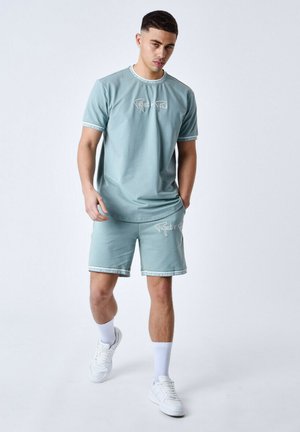 T-shirt en coton bleu clair et short assorti, avec du texte blanc et une finition à motifs sur les manches et l'ourlet, stylé avec des baskets blanches.