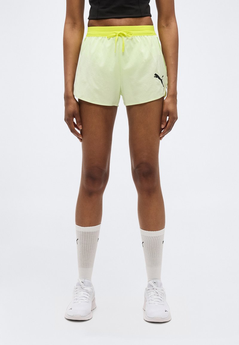 Γυναίκα που φοράει νεον κίτρινο σορτς Puma, λευκές κάλτσες Puma και λευκά αθλητικά παπούτσια Puma, στέκεται μπροστά σε ένα απλό ανοιχτό υπόβαθρο.