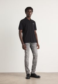 Camiseta polo negra de manga corta con un logo sutil, combinada con jeans gris claro y zapatos negros. Textura suave y diseño ajustado.