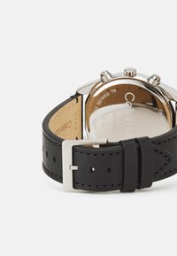 Bracelet de montre en cuir noir avec boucle en argent, présentant un boîtier en acier inoxydable poli et un dos gravé, affichant la marque et les spécifications.