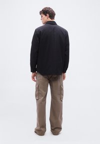 Άνδρας με την πλάτη γυρισμένη φοράει μαύρο μπουφάν και χαλαρά μπεζ παντελόνια cargo, σε απλό λευκό φόντο.