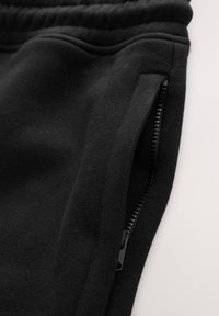 Gros plan d’un pantalon de survêtement en tissu noir montrant une taille élastique et une poche latérale zippée partiellement ouverte.