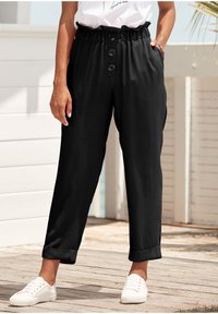 Schwarze weit geschnittene Hosen mit einem elastischen Bund, drei dekorativen Knöpfen, seitlichen Taschen und umgeschlagenen Bündchen, kombiniert mit weißen Sneakers.