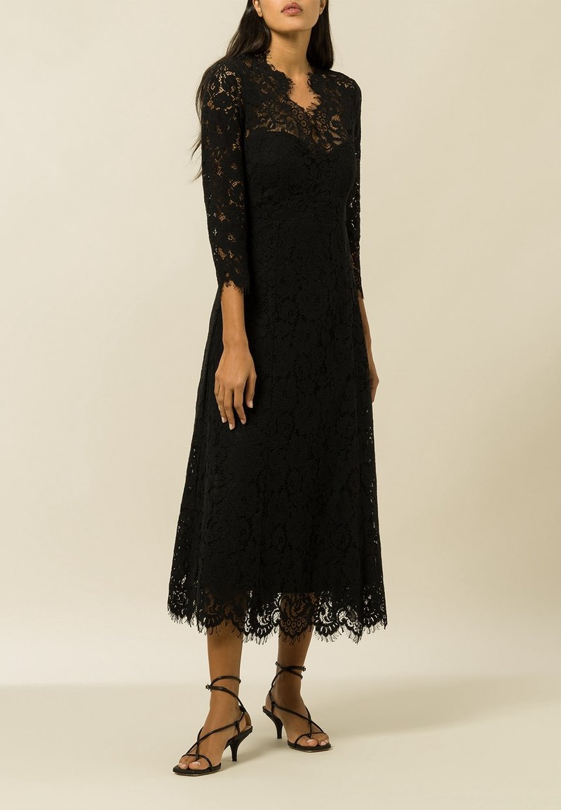 Femme portant une robe midi noire en dentelle à manches longues associée à des sandales à talons noires à brides, sur un fond beige uni.