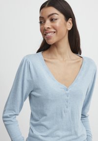 Pull bleu clair à manches longues avec un col en V, comportant une patte de boutonnage et une texture douce. Les manches ont un léger volume au niveau des épaules.
