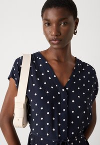 Navy blue dress with white polka dots, v-neckline, short sleeves, and a tied waist. Model holds a cream-colored bag on her shoulder.