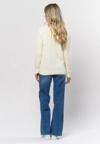 Woman with long blonde hair wearing a cream cable-knit sweater, blue wide-leg jeans, and white sneakers, facing away against a plain background.