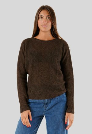 Donna con maglione marrone scuro a maniche lunghe e jeans blu, in piedi davanti a uno sfondo grigio chiaro uniforme.