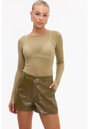 Femme aux cheveux blonds tirés en arrière porte un haut transparent à manches longues olive et un short brillant taille haute olive, une main dans la poche.