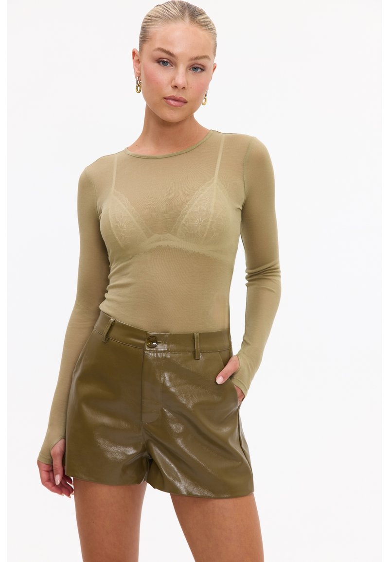 Femme aux cheveux blonds tirés en arrière porte un haut transparent à manches longues olive et un short brillant taille haute olive, une main dans la poche.