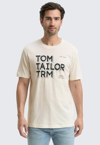 Vīrietis ar tumšiem matiem un bārdu, tērpies krēmkrāsas T-kreklā ar melnu uzrakstu "Tom Tailor TRM" un zilās džinsās, stāv gaismas pelēkā fona priekšā.
