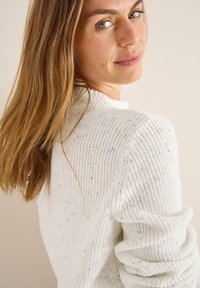 Donna con lunghi capelli castano chiaro che indossa un maglione di maglia bianco, che guarda oltre la spalla con uno sfondo neutro.