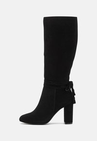 Botas de ante negras hasta la rodilla con punta afilada, tacón grueso y detalles atados en el tobillo. Textura suave con un diseño elegante.
