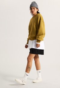 Olijfgroene oversized sweatshirt, witte gelaagde shirt, zwarte shorts, witte sneakers, grijze beanie, zwarte tatoeage op het been, geribbelde witte sokken.
