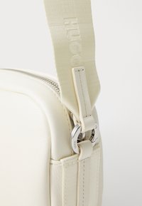 White leather bag with a smooth texture, featuring a silver zipper and hardware. Adjustable fabric strap with logo detail.